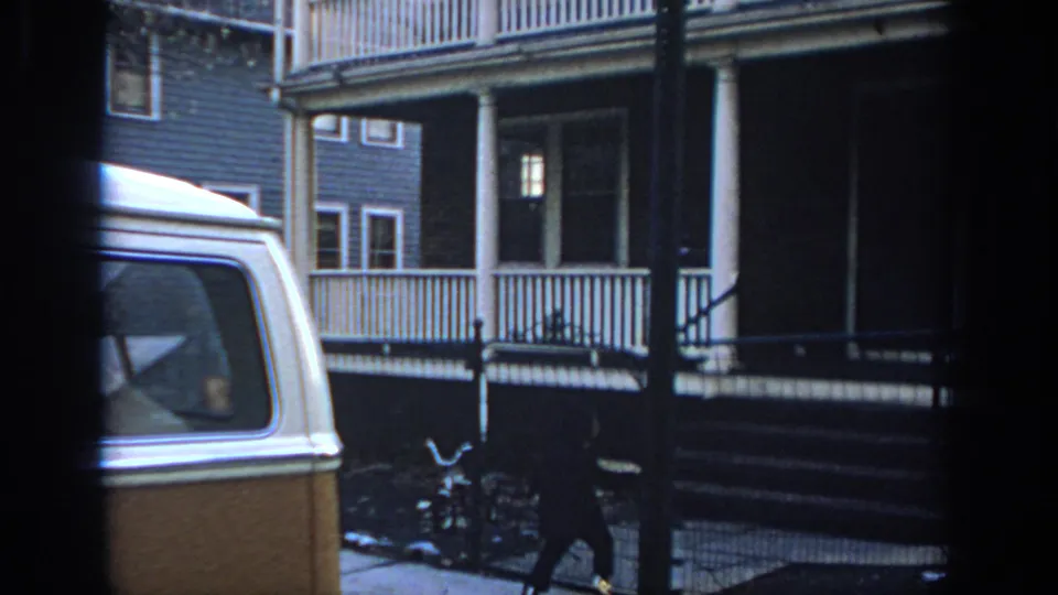 A person passes the front fence of a Philadelphia house, with a two-tone van and porch columns visible.