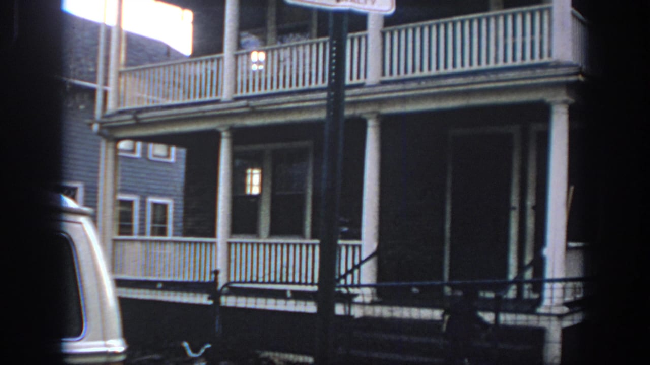 1972 Philadelphia home-movie frame showing a two-story house with stacked white porches, railings, columns, and a partial sign pole in front.