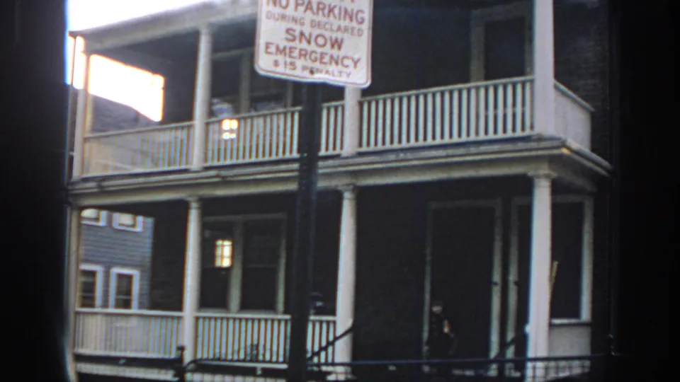 1972 Philadelphia frame showing a no-parking snow emergency sign, stacked porches, railings, columns, and a small person near the stairs.