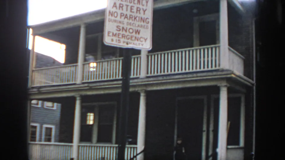 1972 Philadelphia home-movie frame with a snow emergency sign in front of a two-story porch and a small person near the stair railing.