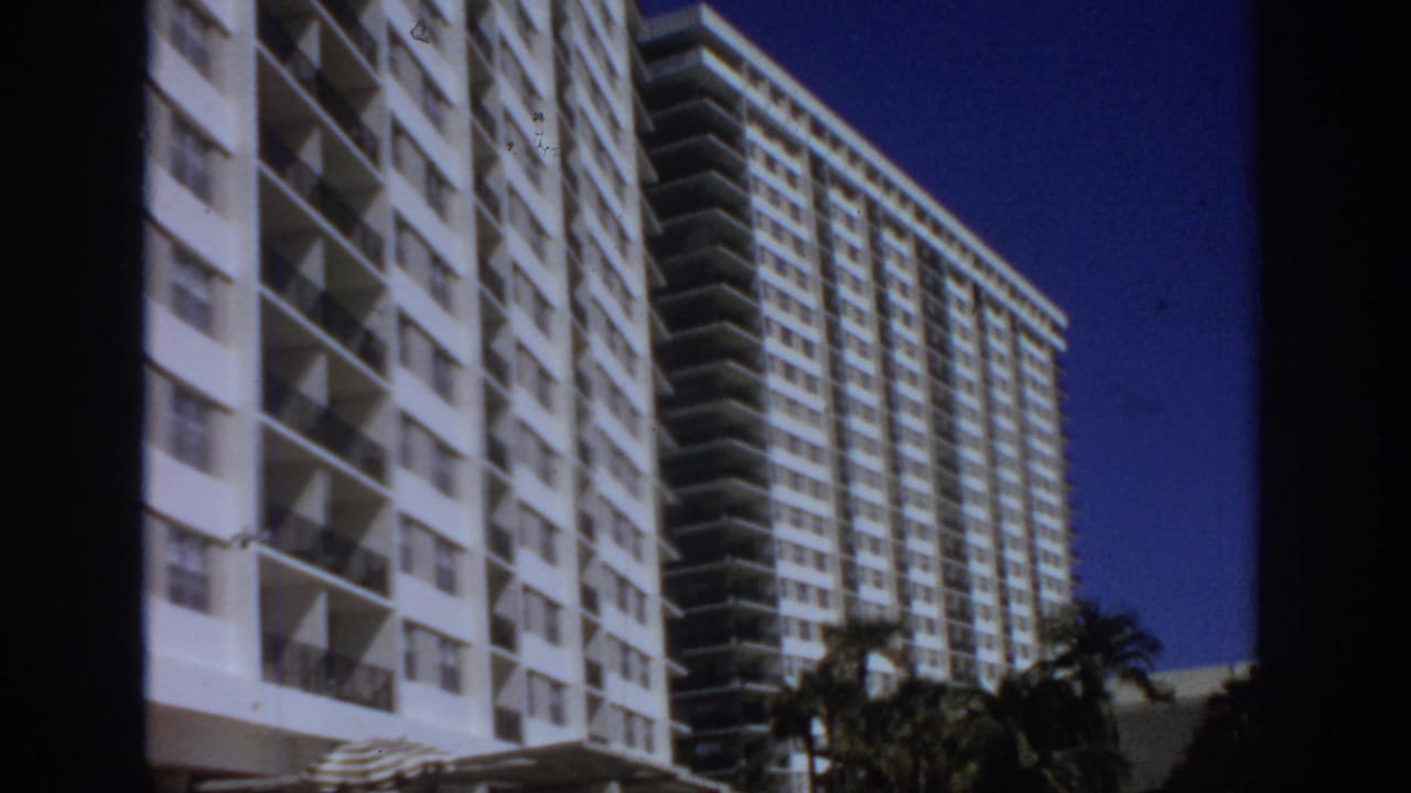 1972 8mm frame of white resort-style hotel towers with repeating balconies, palm trees, and a deep blue sky.