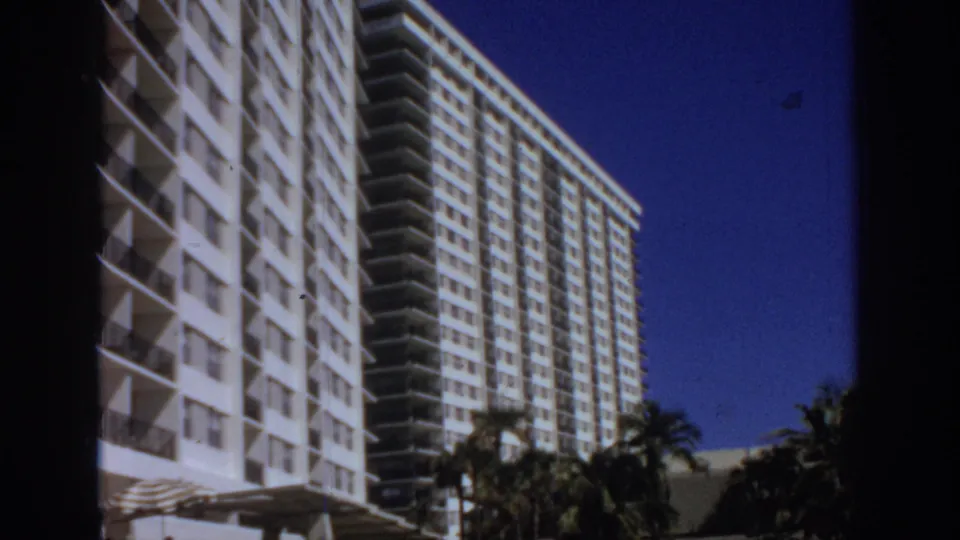 1972 8mm view of a white hotel or resort tower with balconies, neighboring high-rise structure, and palm trees at the base.