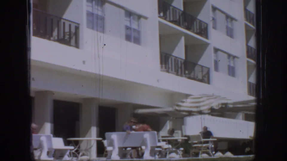 1972 8mm frame showing guests seated at outdoor tables beneath striped umbrellas outside a white hotel building.