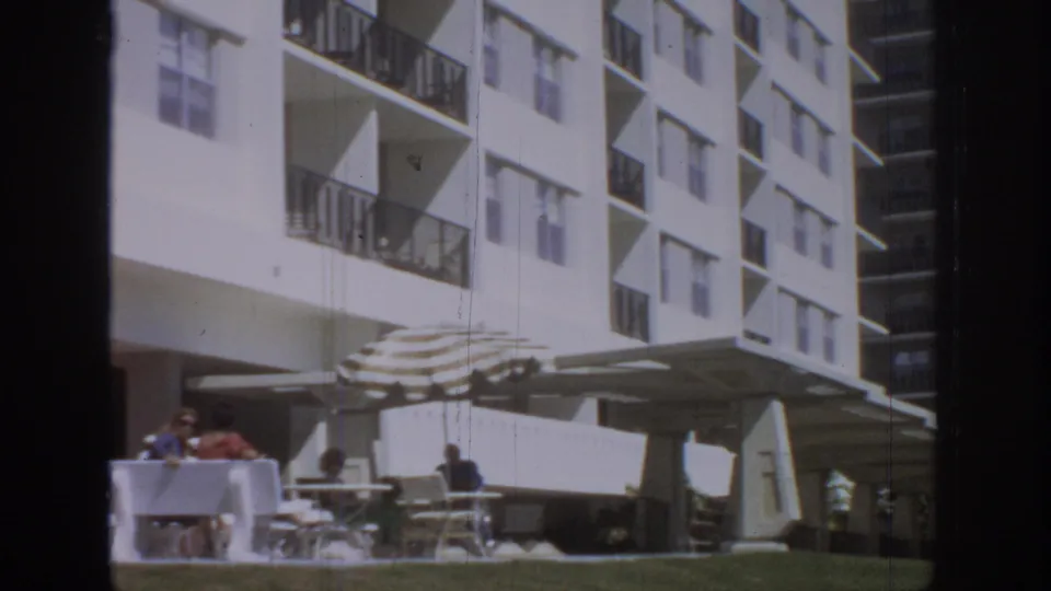 1972 8mm view of a hotel patio where distant guests sit at tables under striped umbrellas beside a white balcony facade.