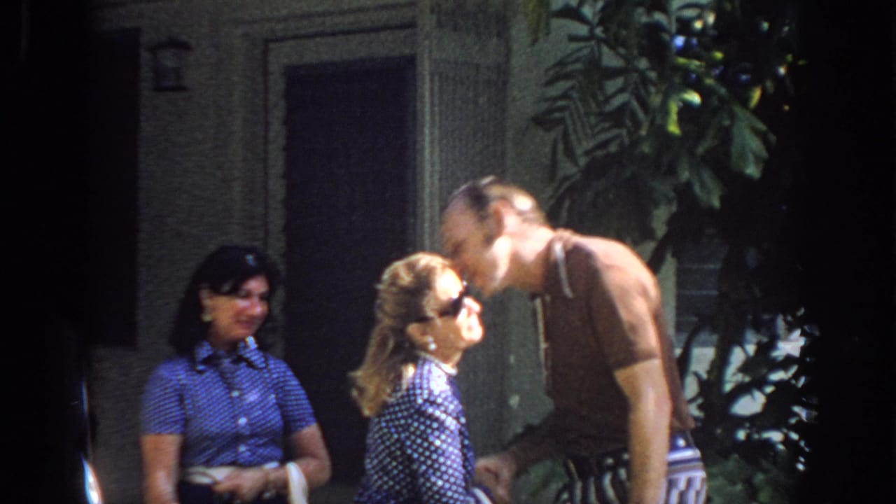 In 1972 Philadelphia home-movie footage, an older man greets a woman with a cheek kiss while another woman watches near a doorway and plants.