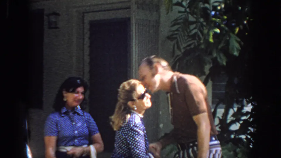 In 1972 Philadelphia home-movie footage, an older man greets a woman with a cheek kiss while another woman watches near a doorway and plants.