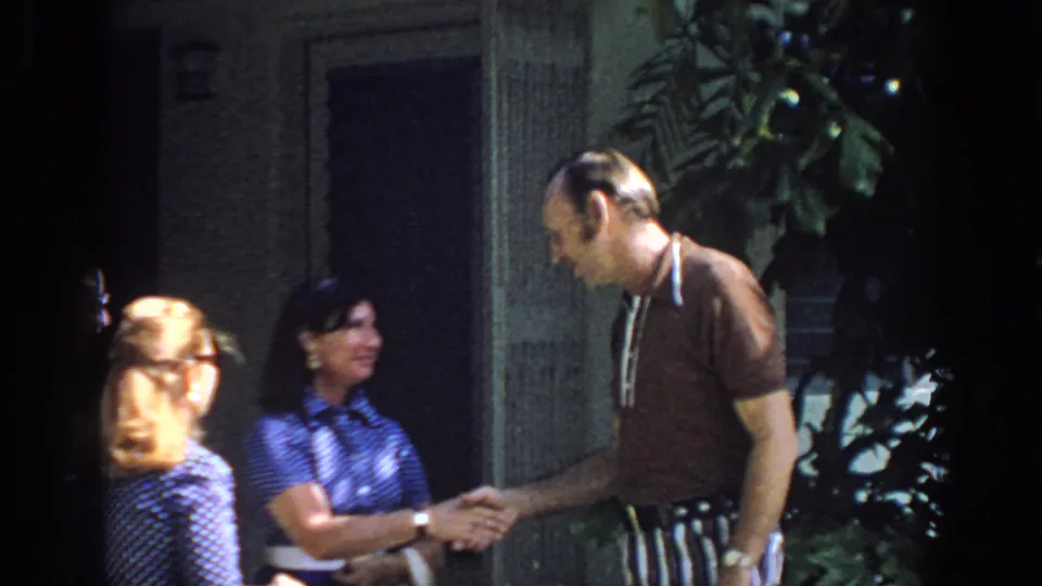 A 1972 Philadelphia home movie frame shows an older man shaking hands with a woman as another woman looks on near an outdoor entrance.