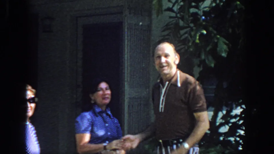 In the 1972 Philadelphia clip, the older man smiles toward the camera as he stands beside women near a doorway and leafy plants.