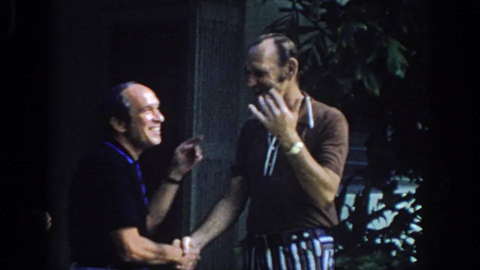 Two adult men in polo shirts share a smiling handshake outdoors in Philadelphia home-movie footage from 1972.