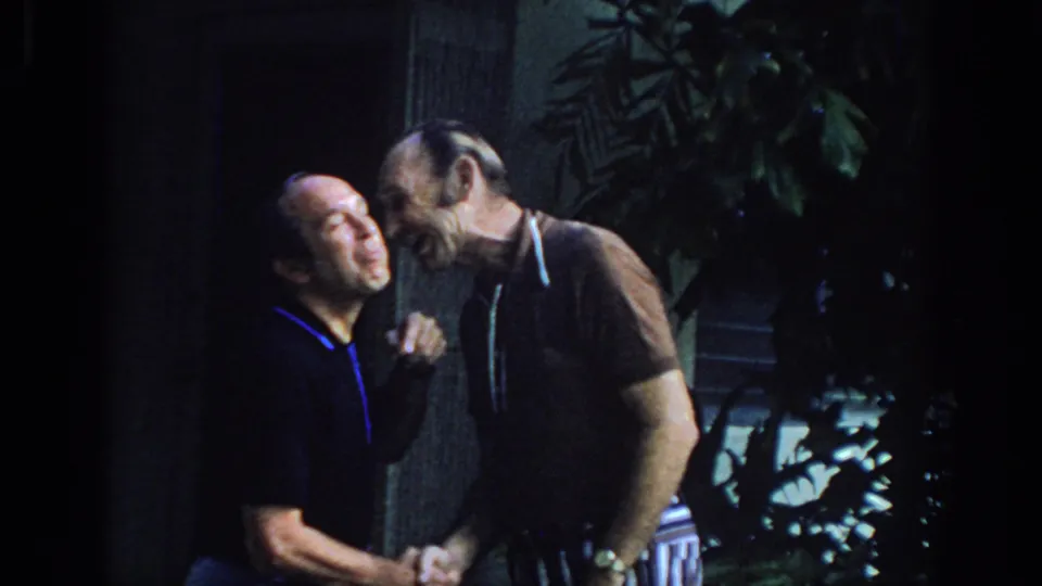 Two adult men laugh together during a handshake in 1972 Philadelphia home-movie footage, with plants and columns behind them.
