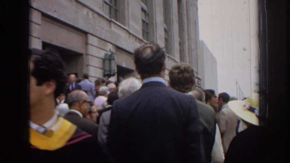 A smiling graduate wearing a yellow stole moves through the foreground as guests and graduates gather outside in Philadelphia in 1972.