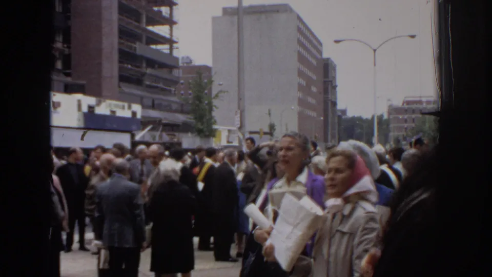 Philadelphia 1972 graduation crowd seen from a handheld 8mm camera, with adults waiting outside city buildings and streetlamps.
