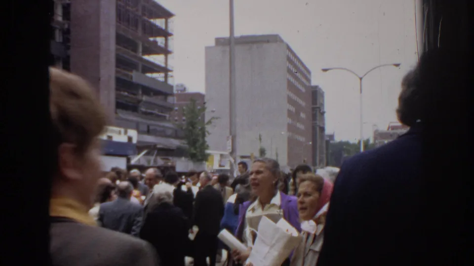 1972 Philadelphia 8mm frame showing graduation attendees on a crowded sidewalk, partly blocked by a close passerby and film-edge shadow.
