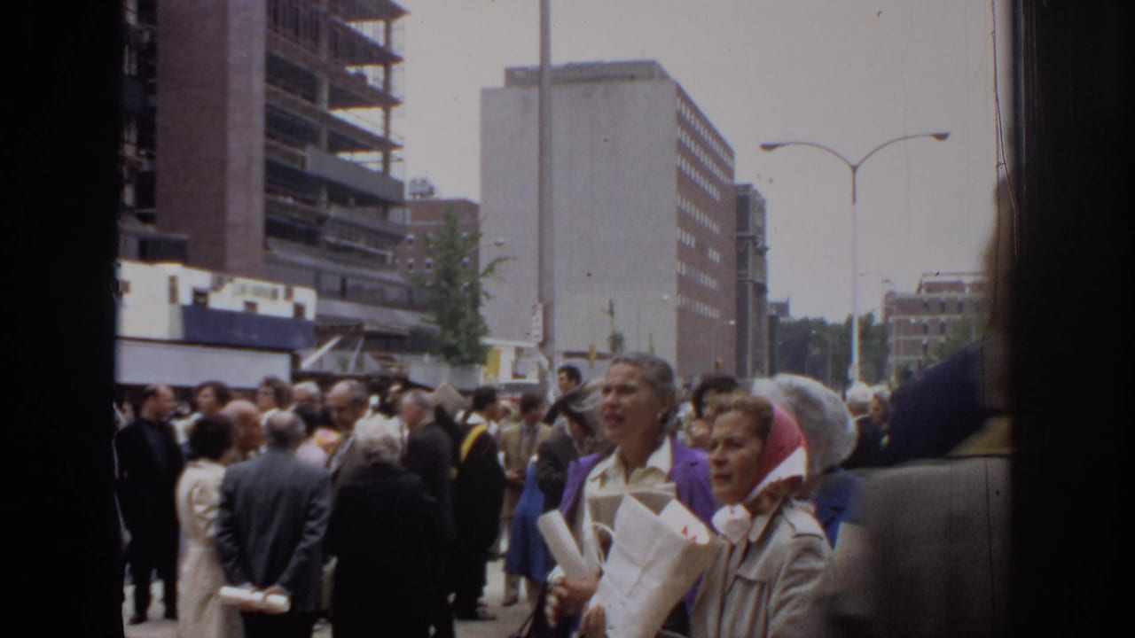 1972 Philadelphia street crowd seen from a handheld 8mm camera, with suited pedestrians, papers, streetlights, and downtown buildings.