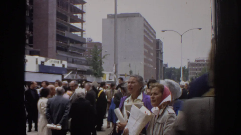 1972 Philadelphia street crowd seen from a handheld 8mm camera, with suited pedestrians, papers, streetlights, and downtown buildings.