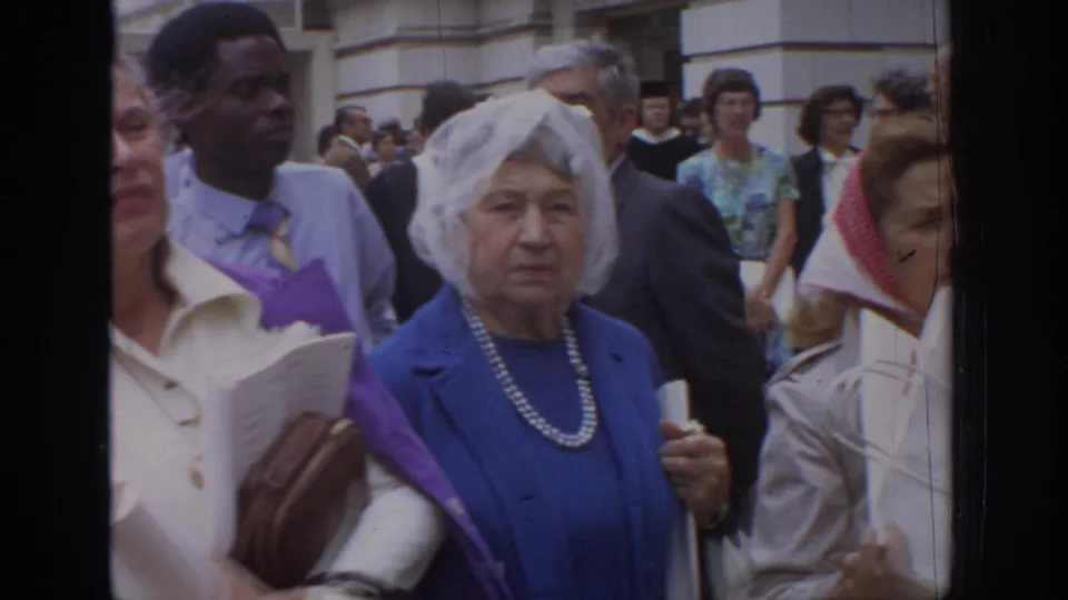 Adults in 1972 Philadelphia street crowd, including an older woman in blue with pearls, people holding papers, and figures in robes behind.