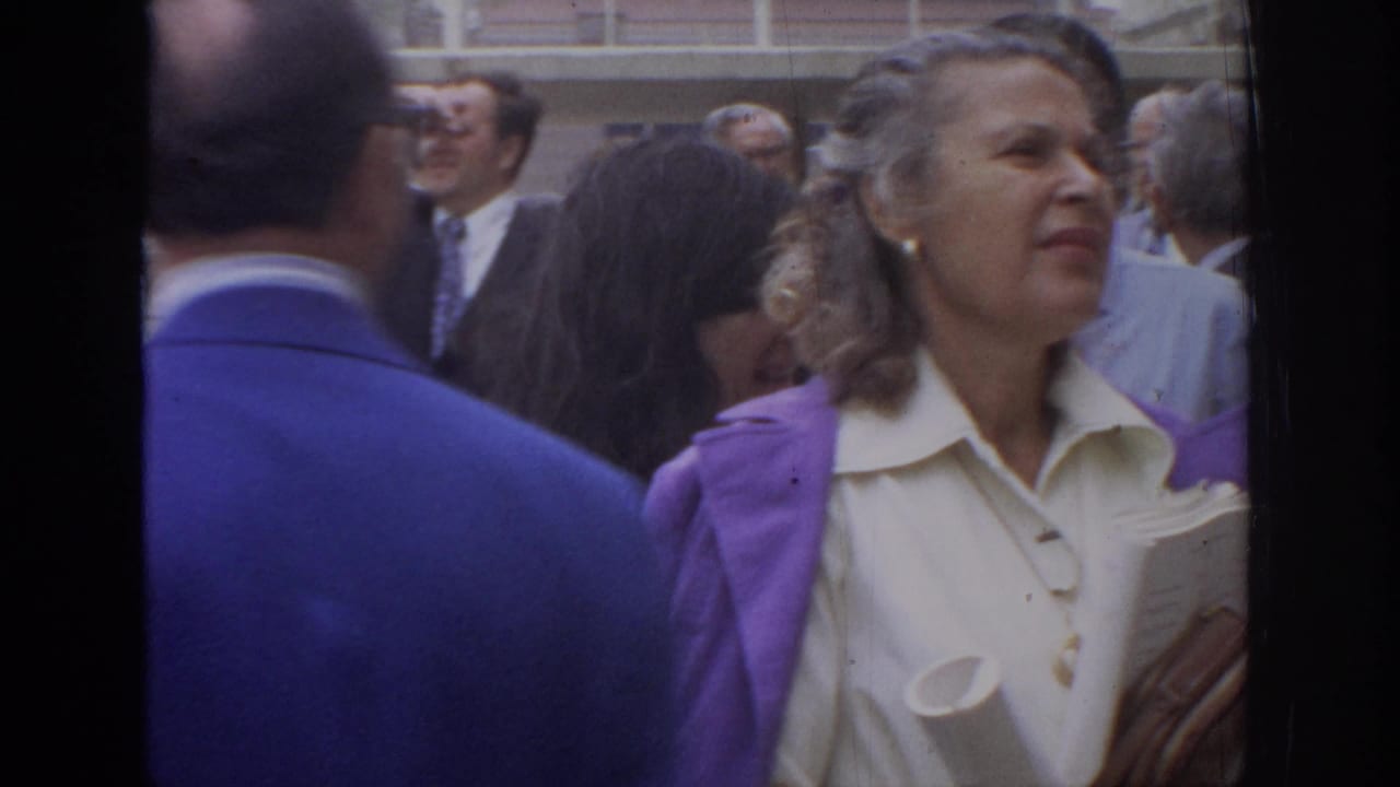 Woman in a white blouse and purple jacket stands among adults in a crowded outdoor area.