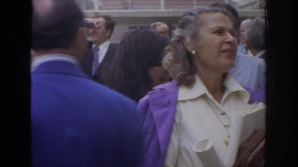 1972 Philadelphia frame with a woman in a white blouse and purple jacket holding papers as adults move around her.