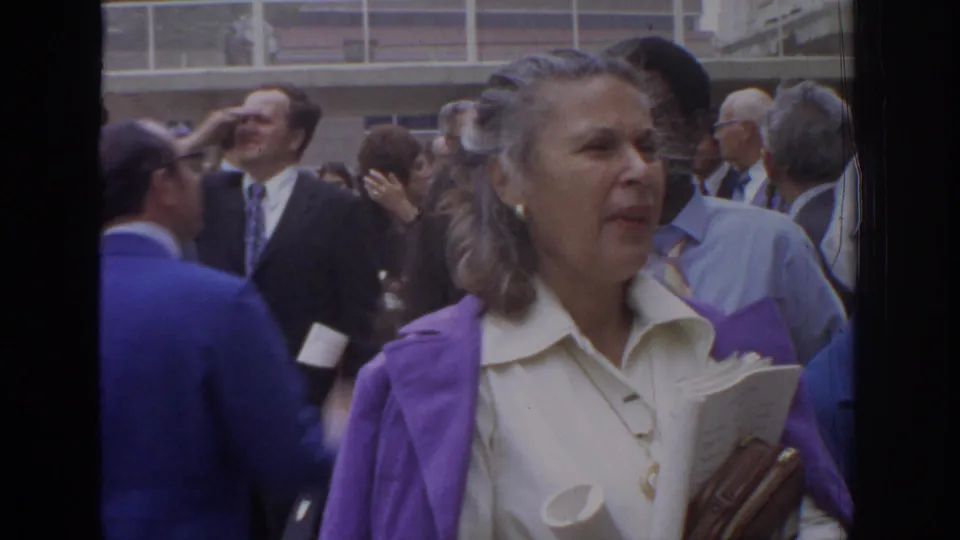 1972 Philadelphia home-movie frame centered on a woman in a purple jacket and white blouse, with suited adults behind her.