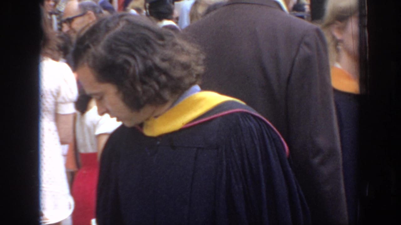 Graduate in black gown looks down while standing in a crowd.