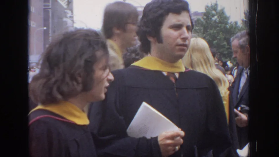Two robed graduates stand in conversation at a 1972 Philadelphia graduation gathering, with one holding papers or a diploma.