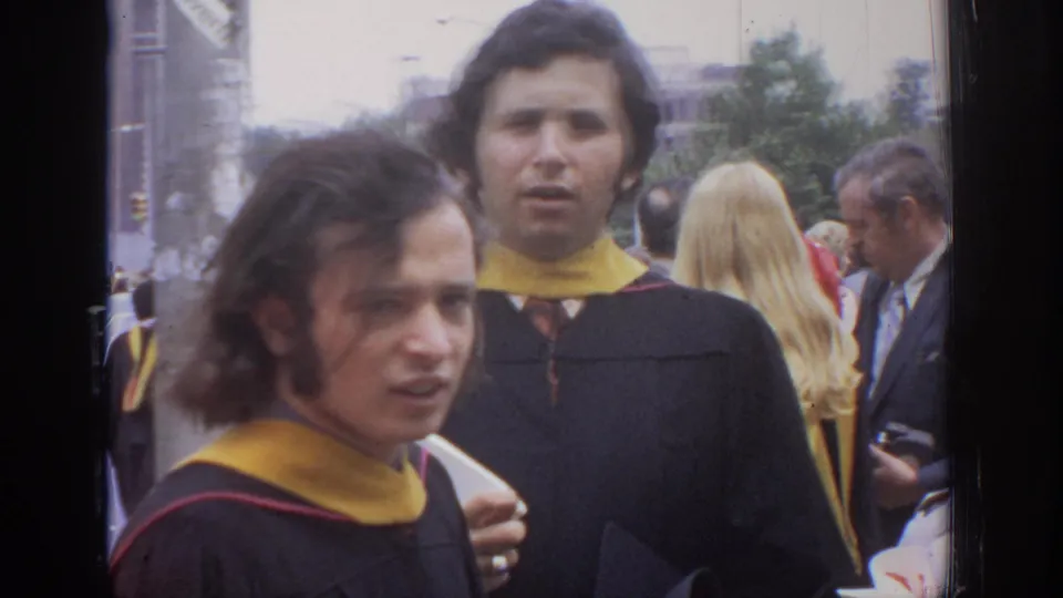 Two graduates in black gowns and yellow hoods face the camera during a crowded 1972 Philadelphia graduation scene.