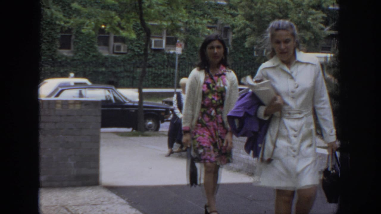 In 1972 Philadelphia footage, two women in period clothing walk along a sidewalk with parked cars and ivy-covered buildings behind them.