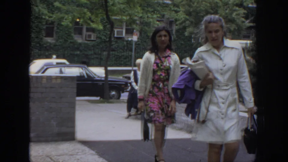 In 1972 Philadelphia footage, two women in period clothing walk along a sidewalk with parked cars and ivy-covered buildings behind them.