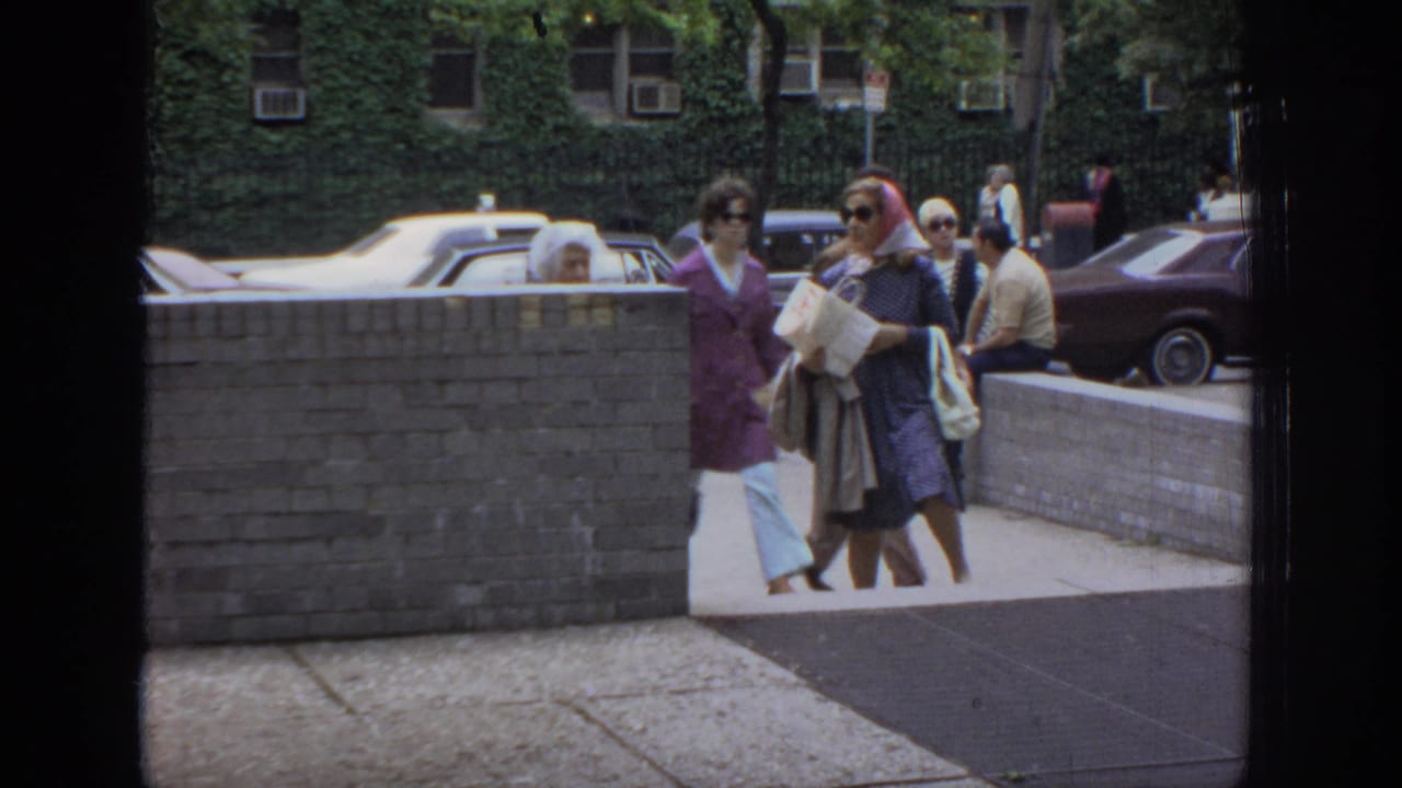 Women in dresses, sunglasses, and headscarves pass along a Philadelphia sidewalk near a brick wall and older parked cars.