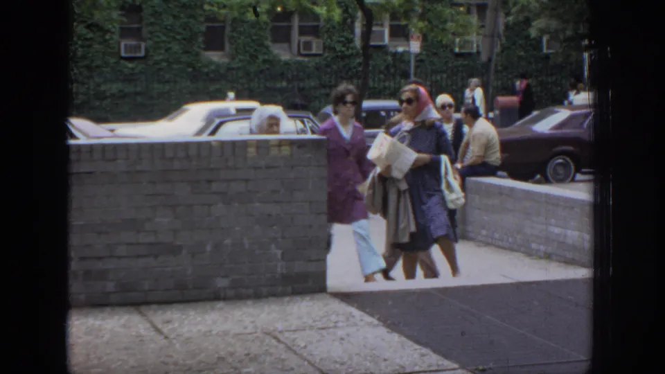 Women in dresses, sunglasses, and headscarves pass along a Philadelphia sidewalk near a brick wall and older parked cars.