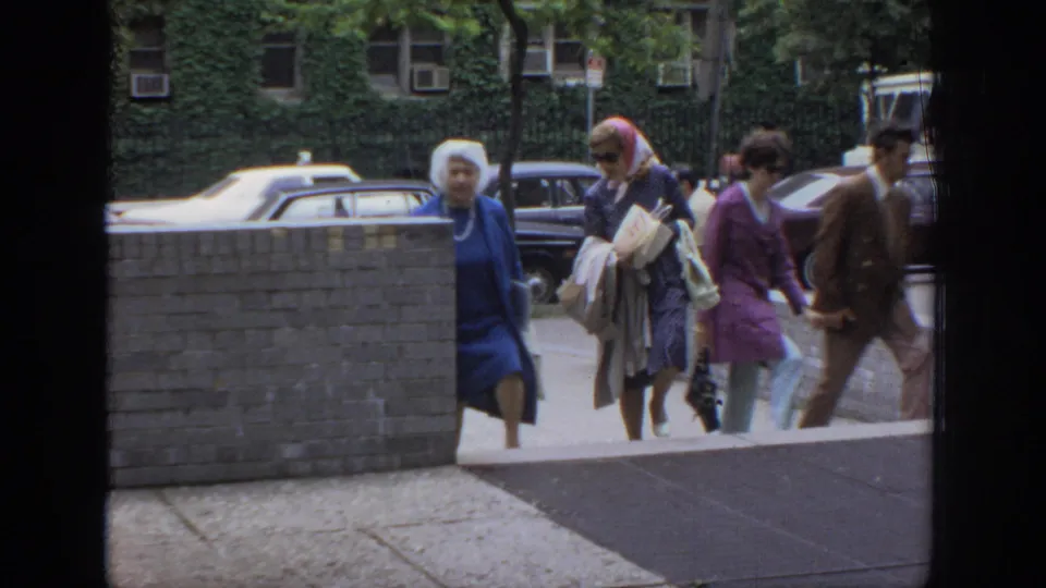 A small group of pedestrians crosses the frame near parked cars and an ivy-covered building in 1972 Philadelphia home-movie footage.