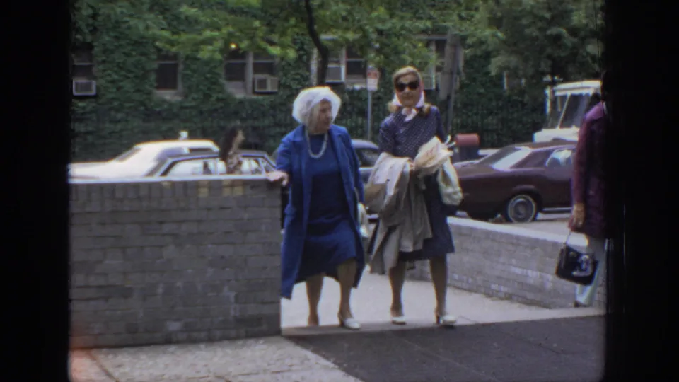 Two women, one in a blue outfit and one carrying folded papers, walk near a low brick wall in the 1972 Philadelphia street scene.