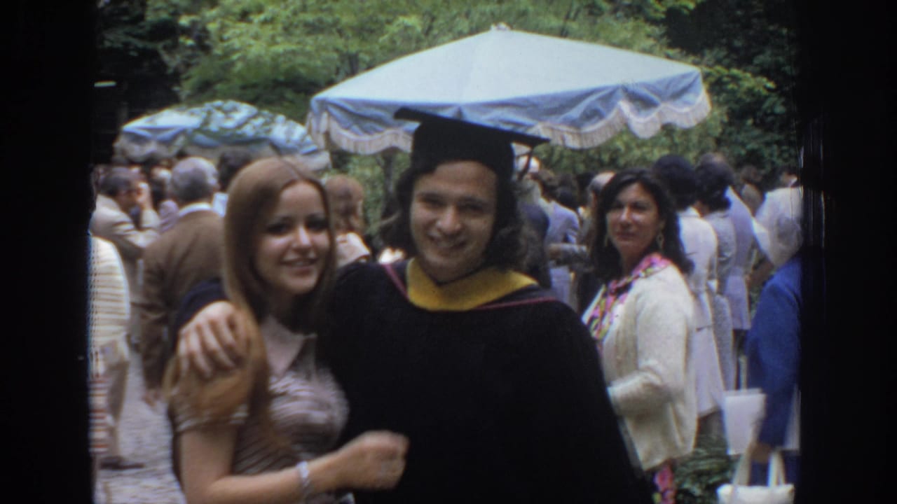 In a 1972 Philadelphia home-movie frame, a young woman poses beside a graduate in cap and gown in a crowded outdoor gathering.