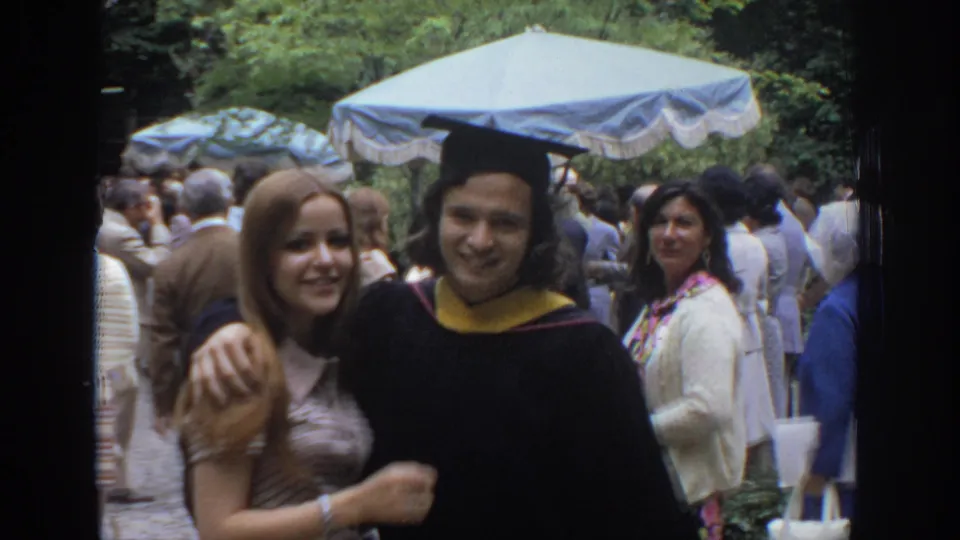 In a 1972 Philadelphia home-movie frame, a young woman poses beside a graduate in cap and gown in a crowded outdoor gathering.
