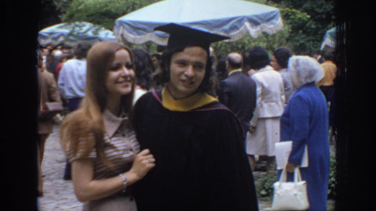 Graduate in cap and gown walks beside a young woman in a striped dress at an outdoor event.