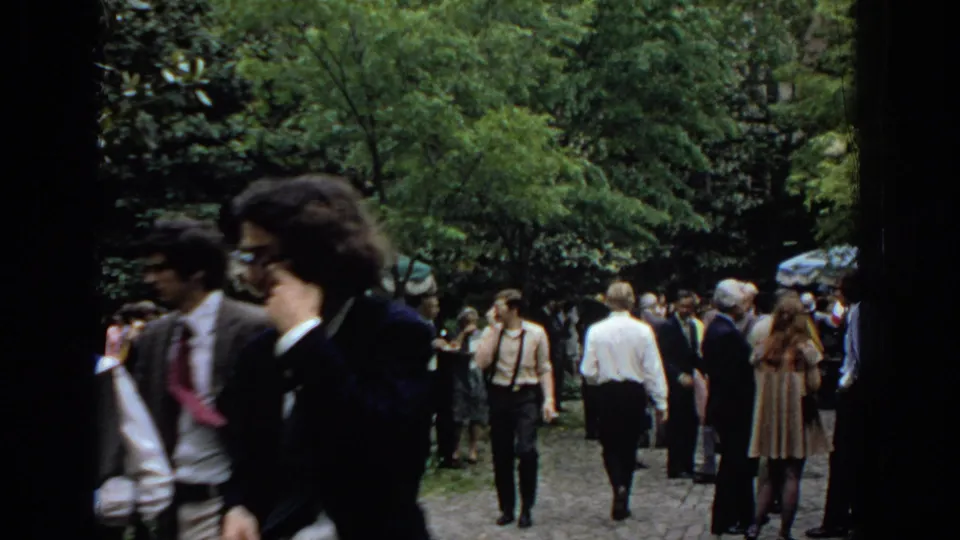 The home-movie view shifts to the surrounding graduation crowd, with formally dressed guests moving through a leafy outdoor space.