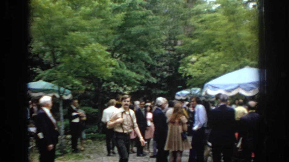 A wide 1972 Philadelphia graduation crowd view shows guests under umbrellas and trees, with a man walking toward the camera.