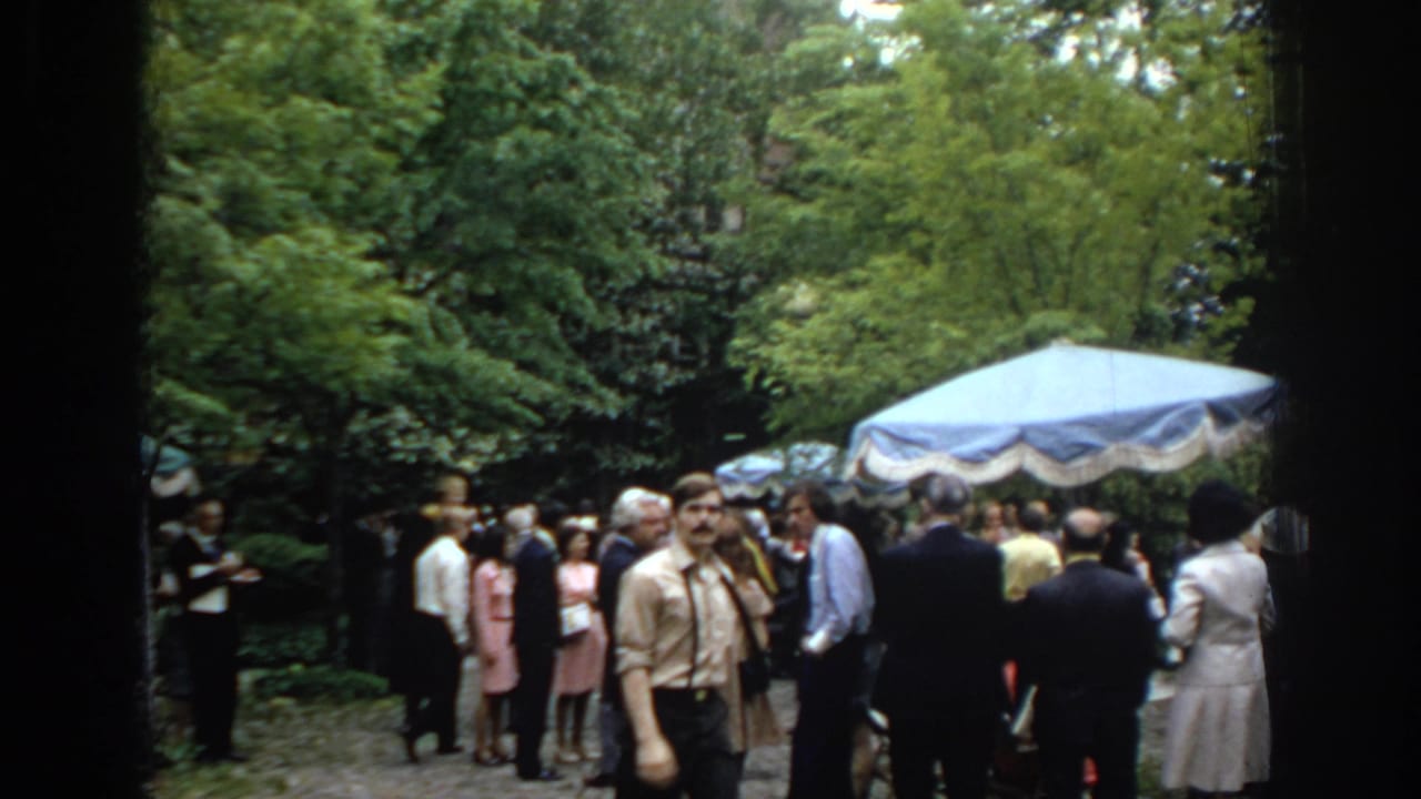 1972 Philadelphia home-movie frame showing adults in period clothing gathered near a blue canopy umbrella in a tree-lined garden.