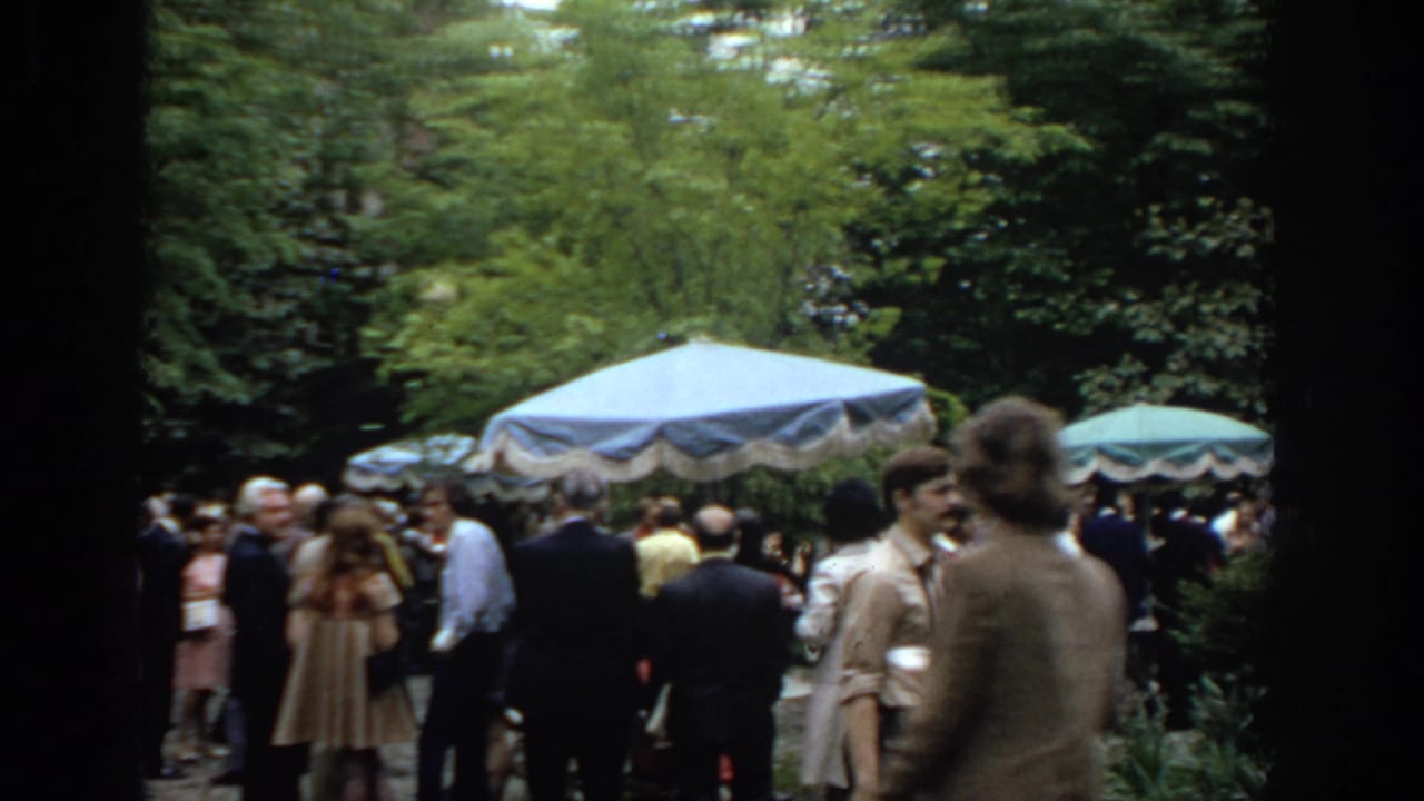 Crowd stands under trees beside blue and green umbrellas.