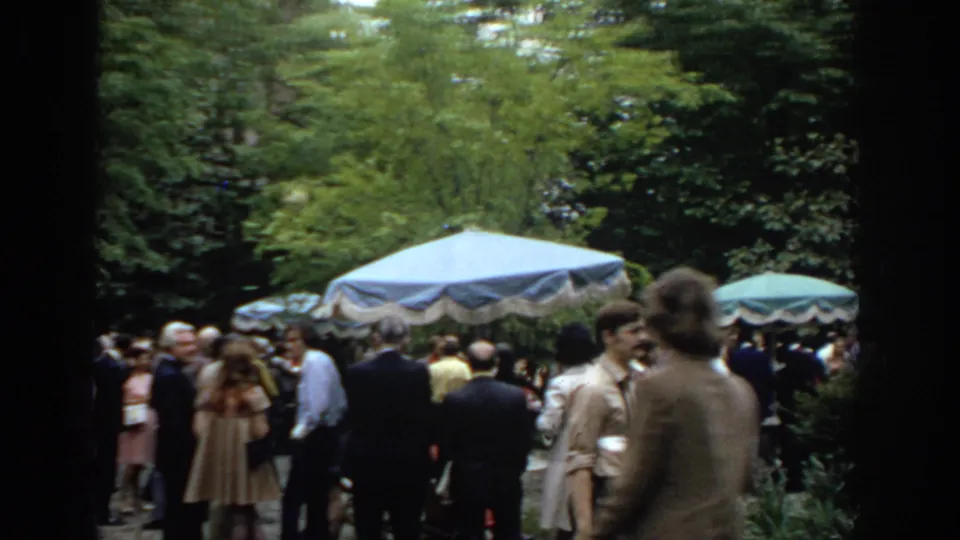 Adults in 1970s attire stand and talk beneath trees beside blue and green umbrellas in the Philadelphia garden scene.