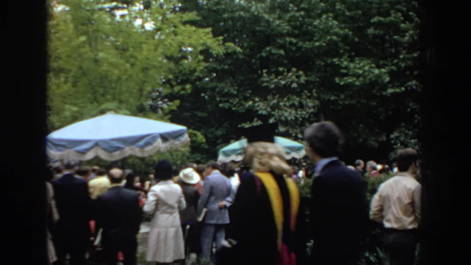 A wider 1972 Philadelphia home-movie view shows adults gathered around a blue umbrella, with trees and garden foliage behind them.