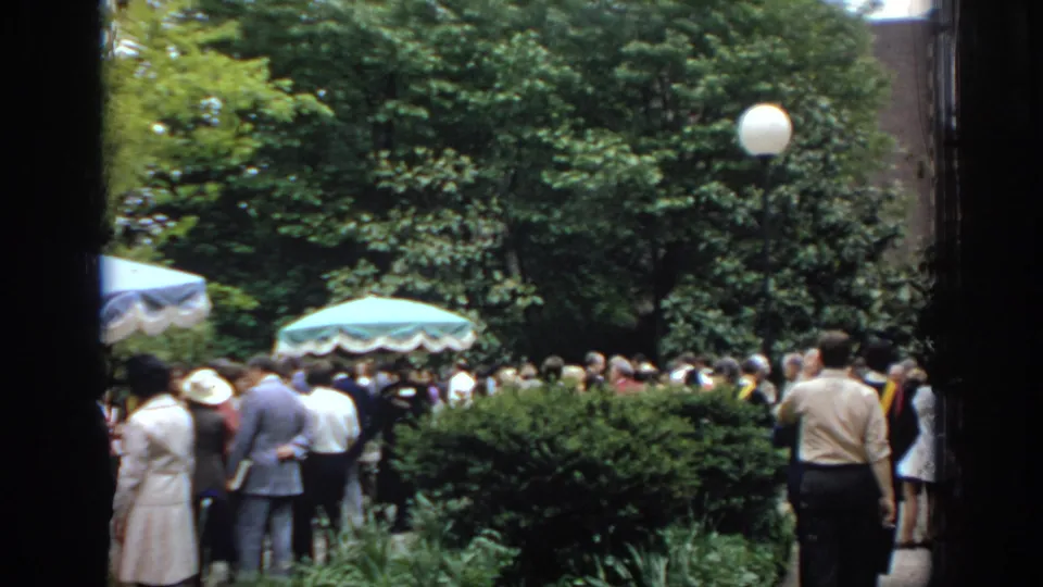 Adults viewed from behind gather in a leafy Philadelphia garden, with canopy umbrellas, shrubs, and a round globe lamp visible.
