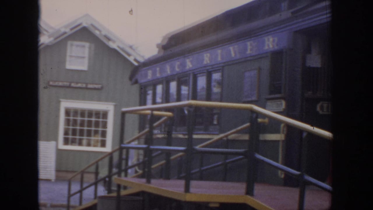 1972 home-movie frame of a dark heritage railroad car beside a green station building and wooden boarding platform in Philadelphia, Pennsylvania.