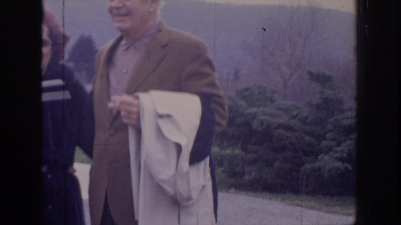 Adults outdoors, including a man in a brown jacket holding a light coat.