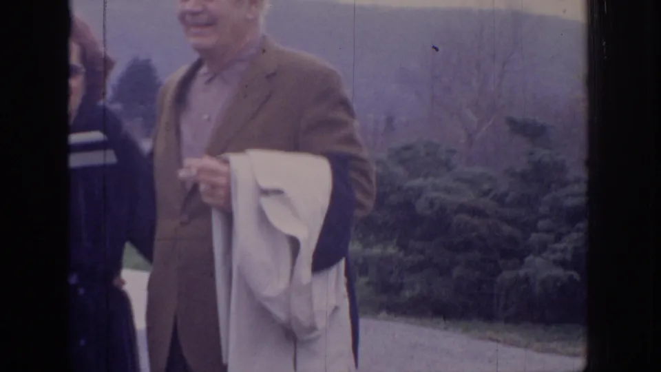 1972 Philadelphia home-movie frame with adults outdoors; a man in a brown jacket holds a light coat near shrubs and distant hills.