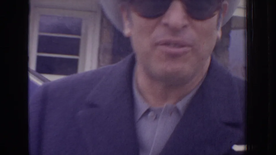 Close 8mm frame from the 1972 Philadelphia clip showing an adult man in dark sunglasses and a coat near a building.