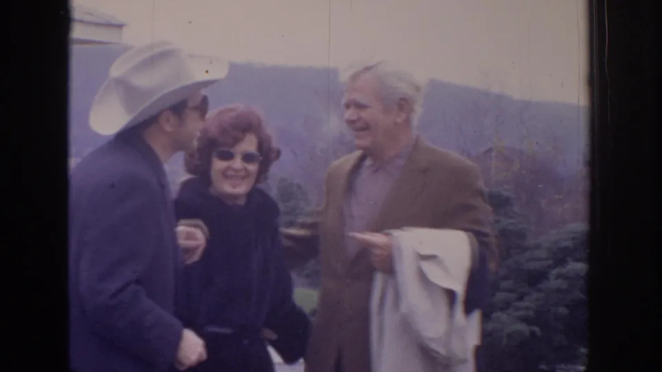 1972 Philadelphia home-movie frame showing three adults talking and smiling outdoors, including a man in a cowboy hat and a man holding a coat.