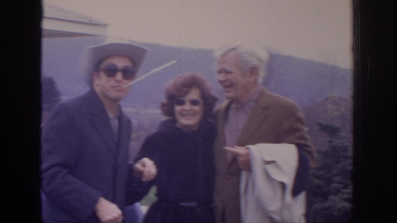 1972 home-movie frame of three adults outdoors near hills, including a man in a light cowboy-style hat, a red-haired woman, and an older man in a blazer.