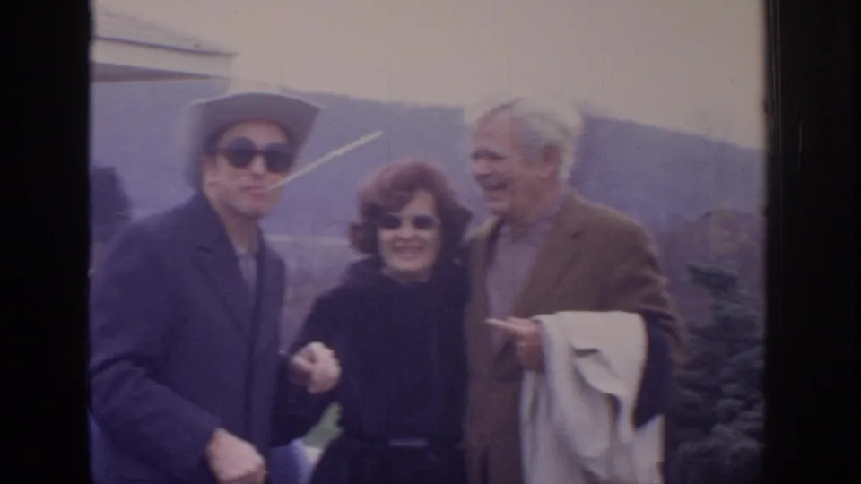 1972 home-movie frame of three adults outdoors near hills, including a man in a light cowboy-style hat, a red-haired woman, and an older man in a blazer.