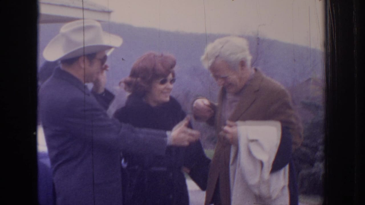 A man in a light hat gestures toward an older man as a woman stands between them outdoors.